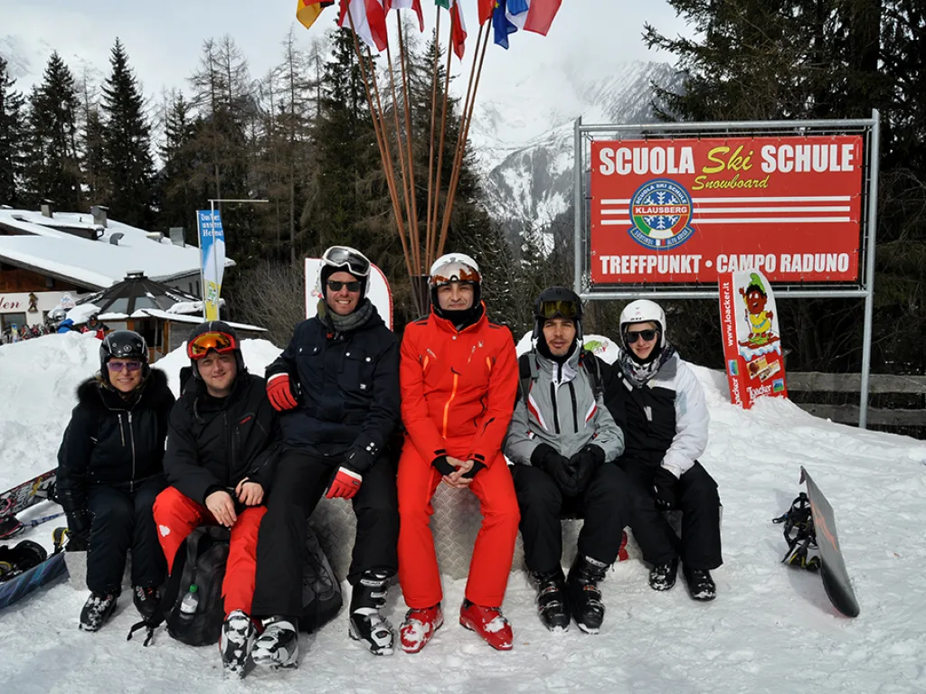 Gruppe sitzt vor dem Schild der Skischule im Schnee.